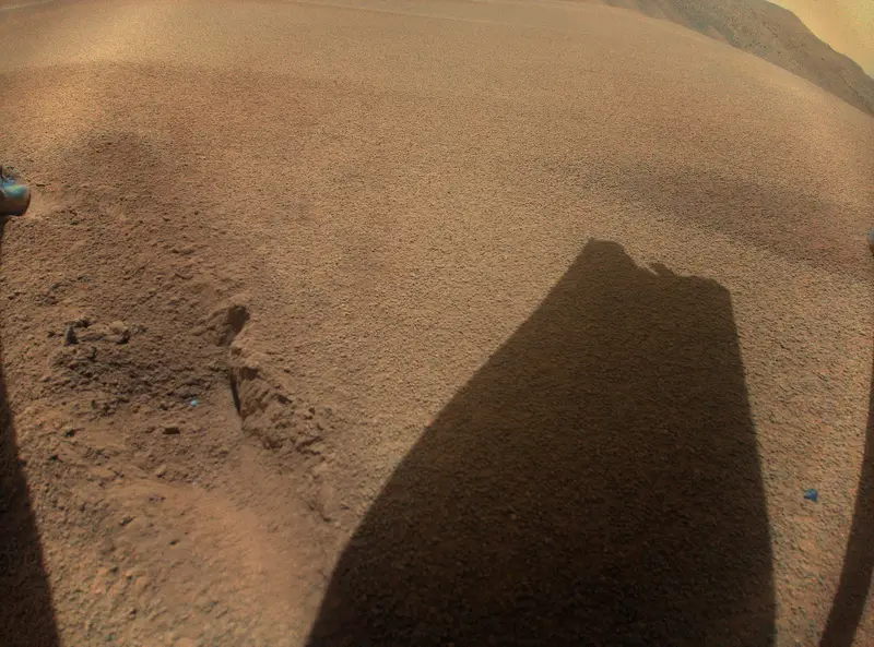 Rippled reddish sand with the shadow of Ingenuity's rotor, one blade visibly shorter